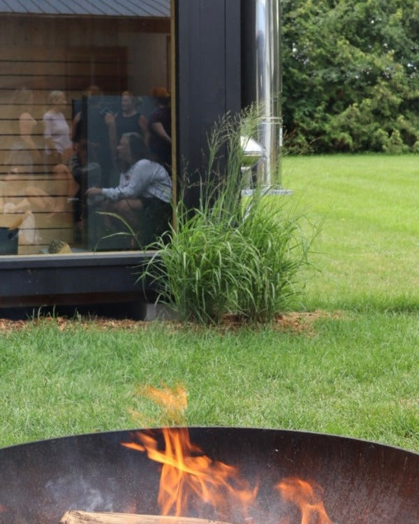 The Sip Saunas Sauna Garden: guests inside the black sauna, seen through its full-height window, fire pit burning in the foreground