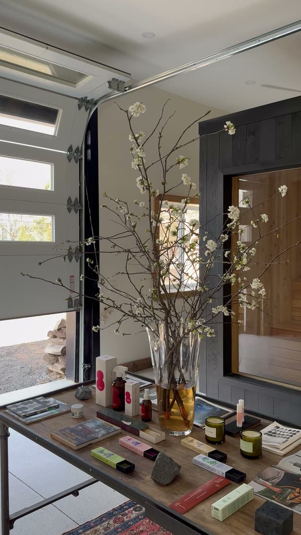 The Sip showroom with the garage door open: blossom branches in a tall vase on the product table, and the black sauna with its window on the right